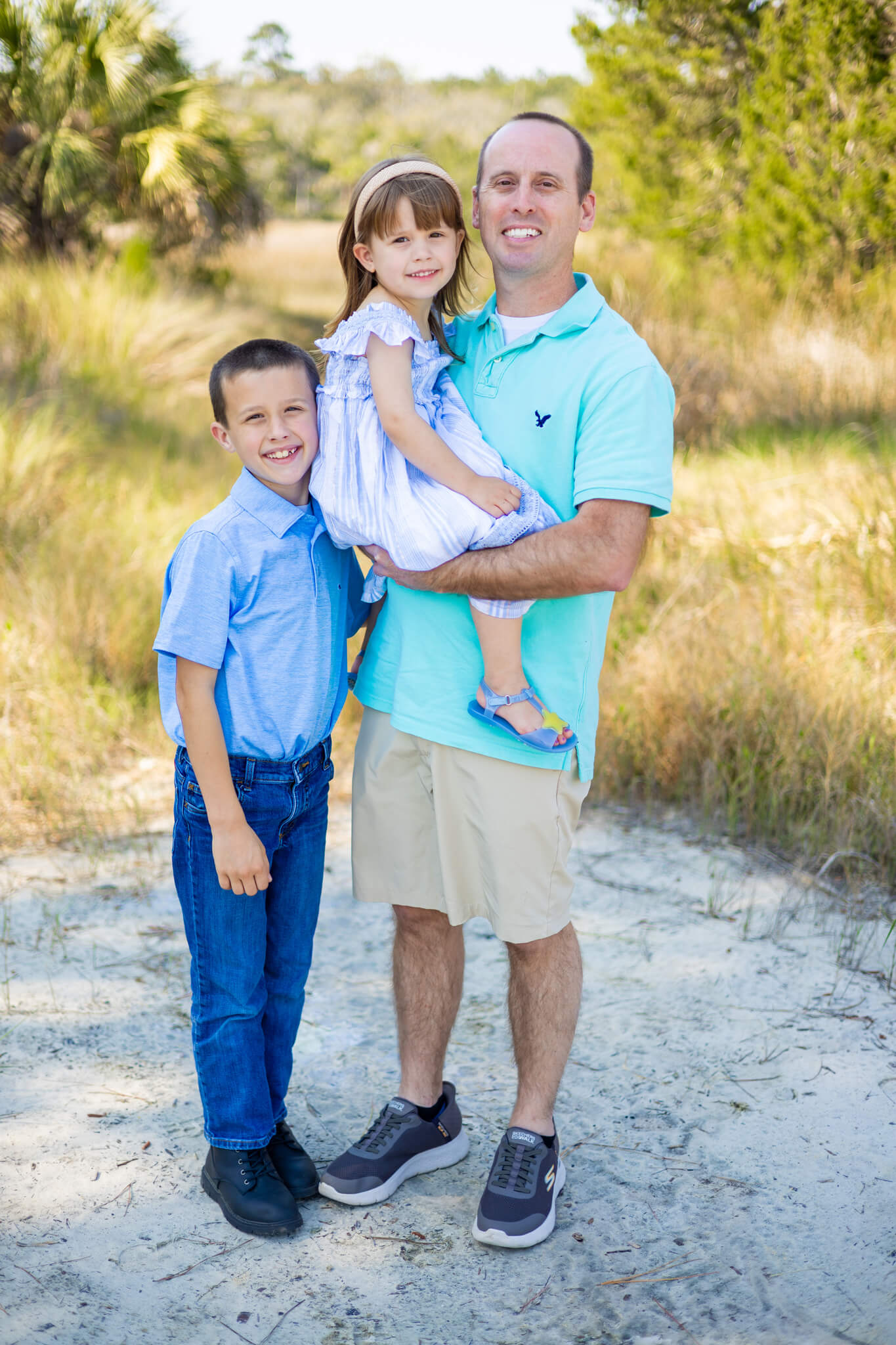 A smiling father in a teal polo stands holding his young daughter as his smiling son in blue stands with them thanks to marriage counseling in richmond hill, GA