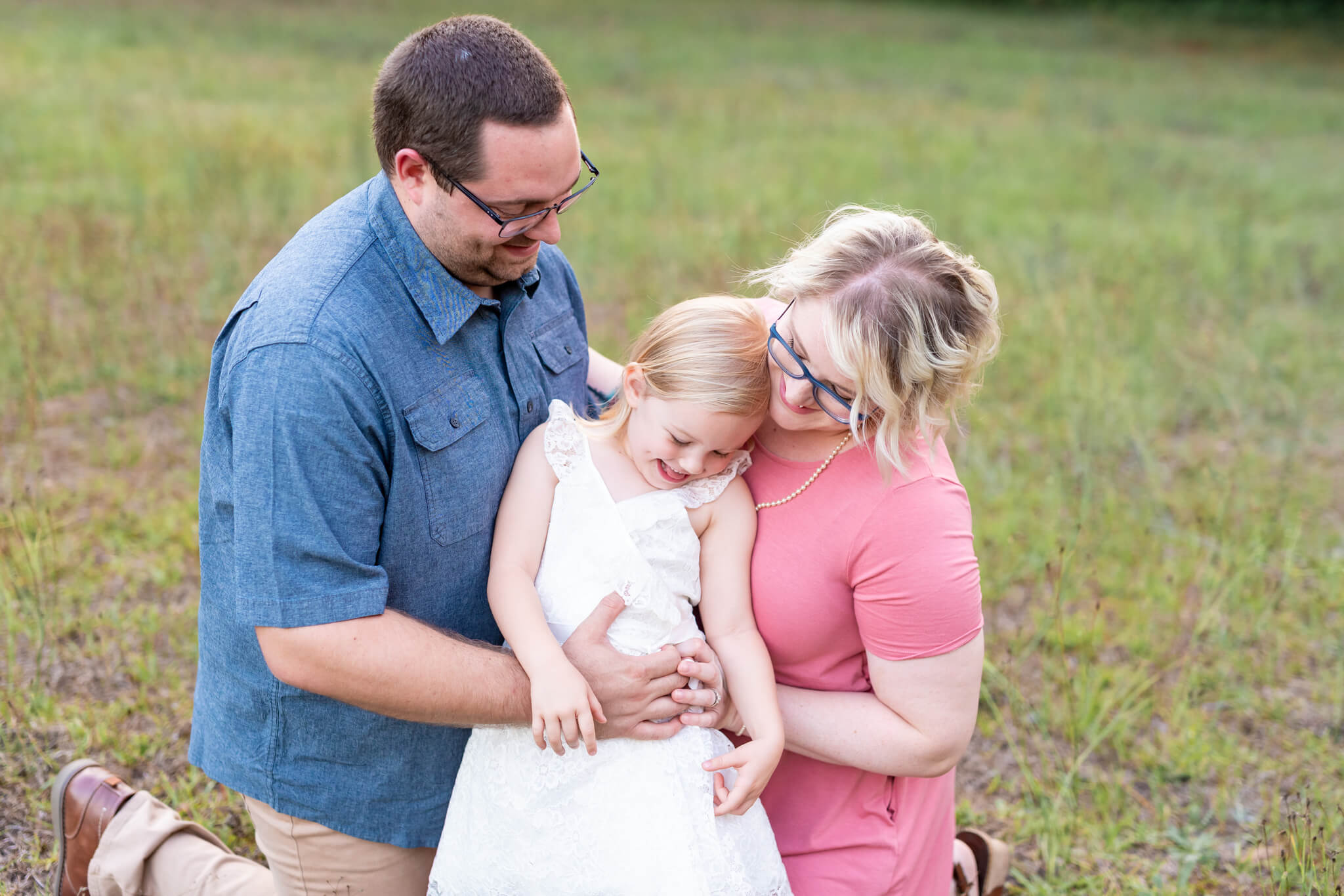 A mom and dad play with their young daughter in a park in a white dress