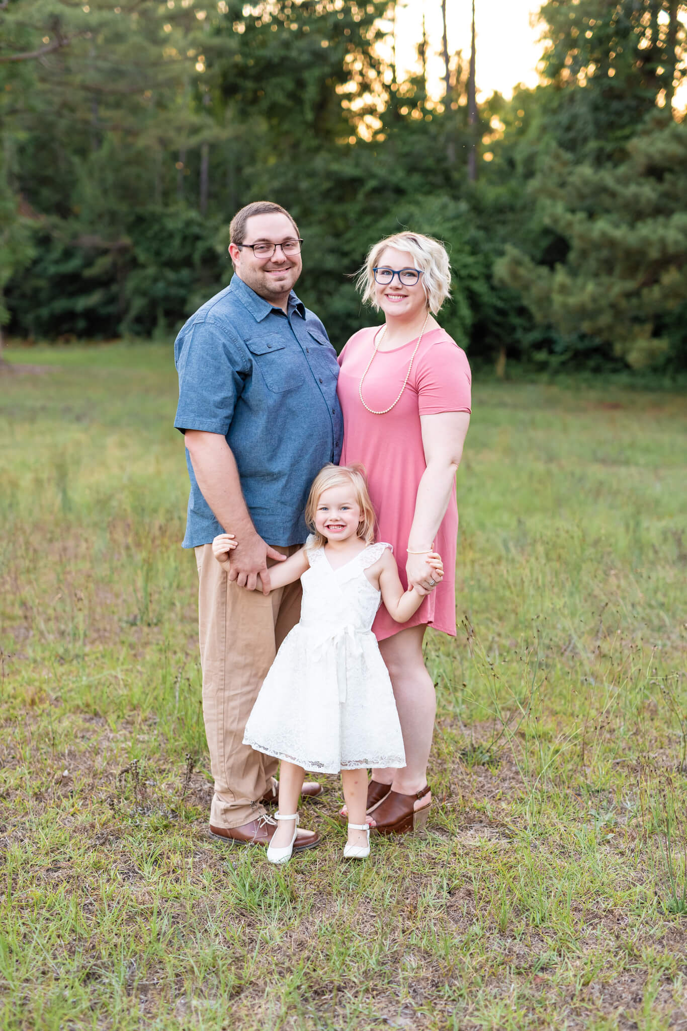 Happy mom and dad in blue and pink stand in a park holding hands with their young daughter before visiting romantic restaurants in richmond hill, GA