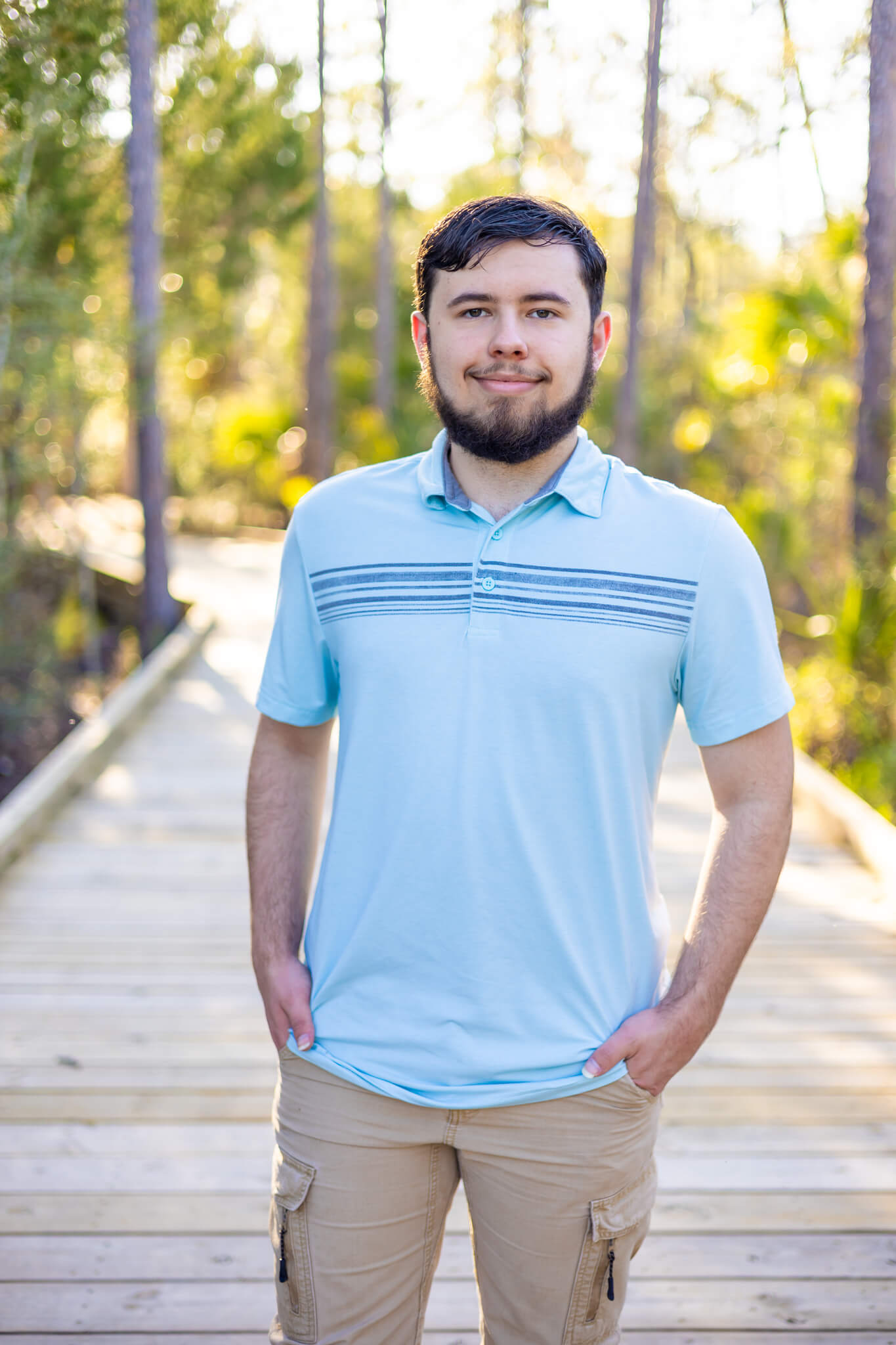 A smiling high school senior in a blue polo walking on a boardwalk at sunset with hands in pockets