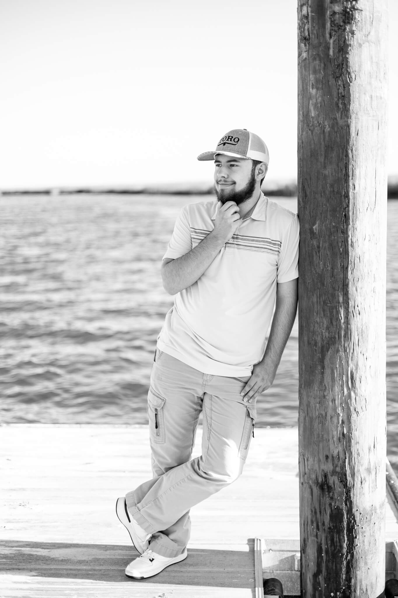 A black and white image of a high school senior in a polo and hat leaning on a dock pilling after some volunteer opportunities in pooler ga