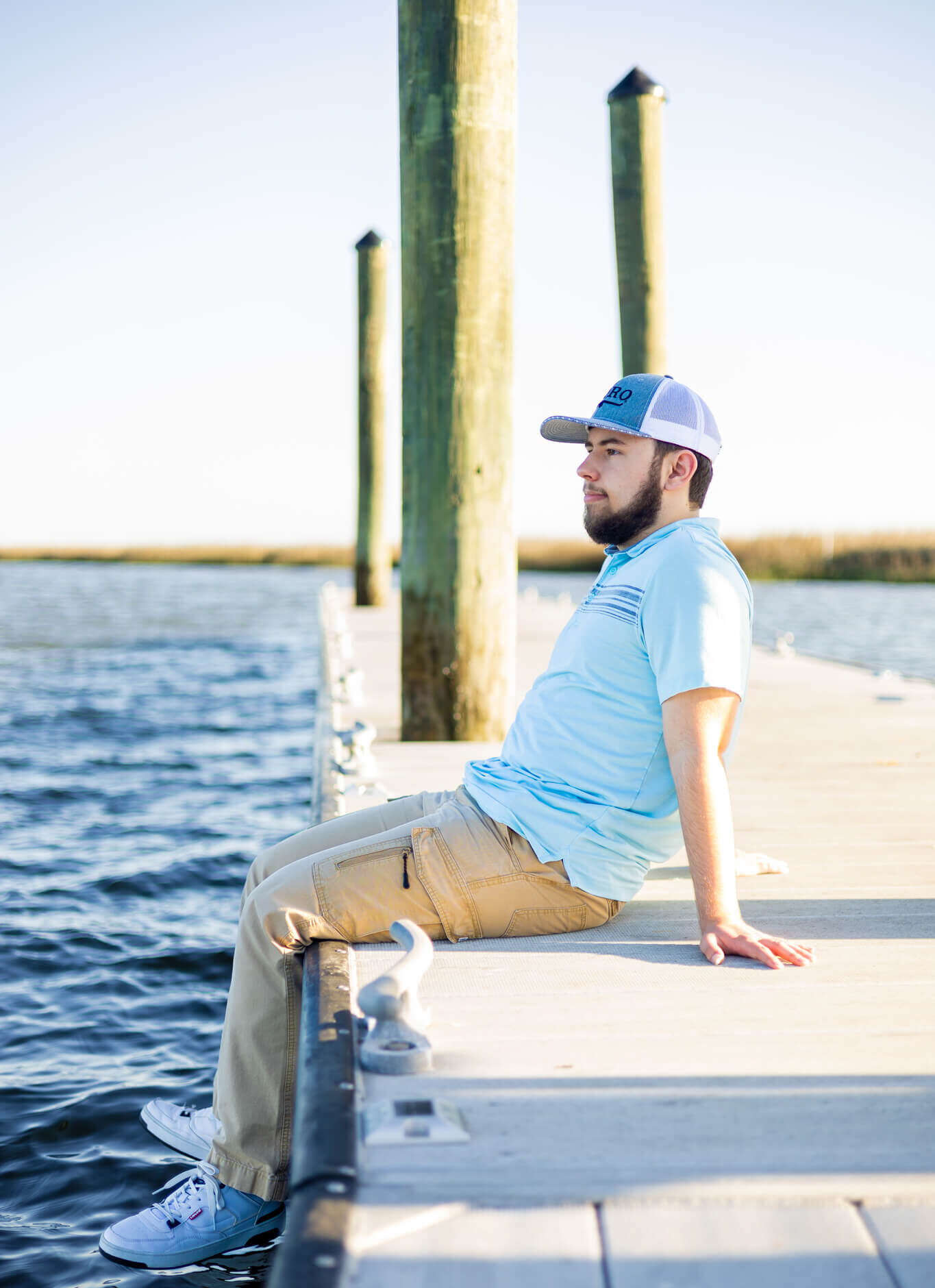 A high school senior in a blue polo and matching hat sits on a dock at sunset after enjoying volunteer opportunities in pooler ga