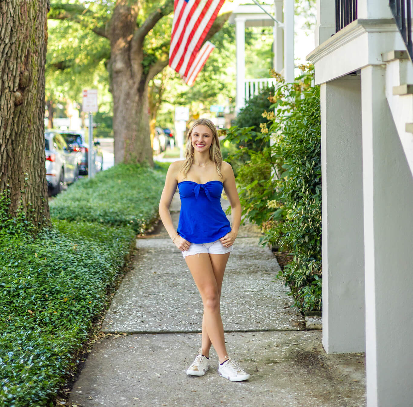 A smiling high school senior in a blue blouse and white shorts stands with thumbs in belt loops on a sidewalk under american flags after shopping boutiques in brunswick ga