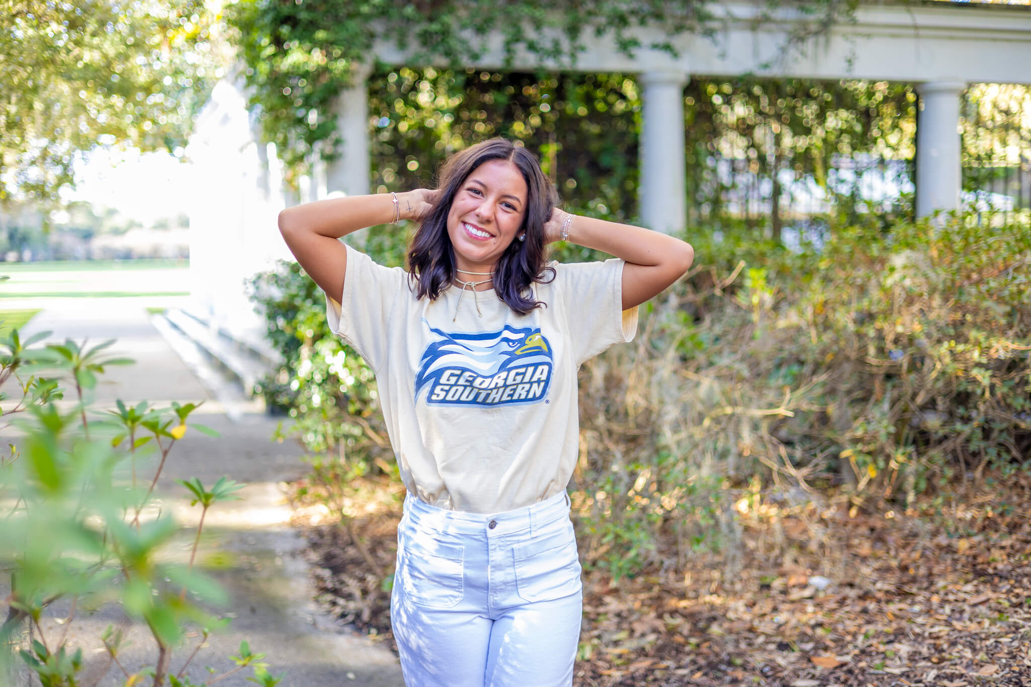 A happy high school senior wearing a shirt of one of the colleges near richmond hill ga walking in a garden witha rms behind her head