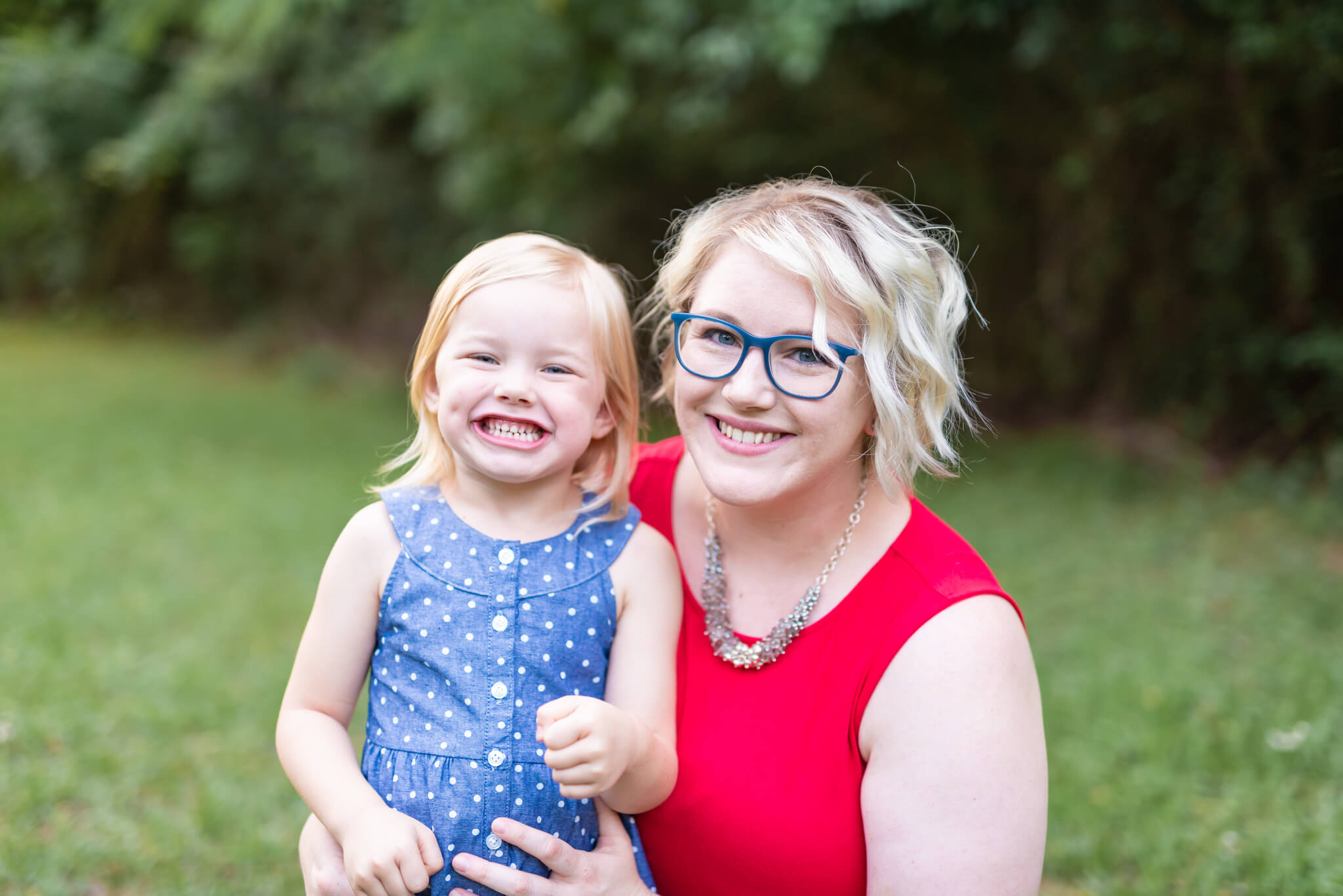A smiling toddler girl in a blue dress sits with mom in red in a park lawn