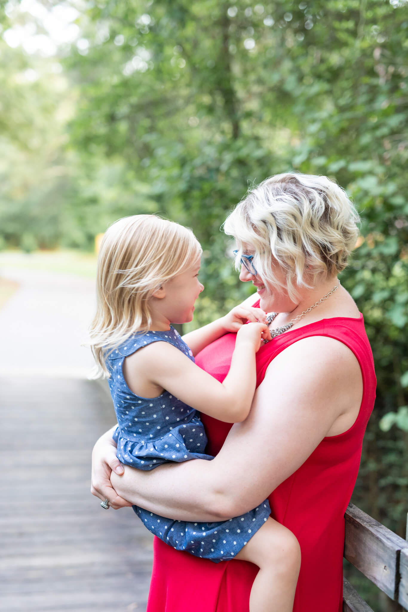 A mom in a red dress plays with her toddler daughter in blue dress in her arms on a park boardwalk after enjoying a couples massage in richmond hill ga