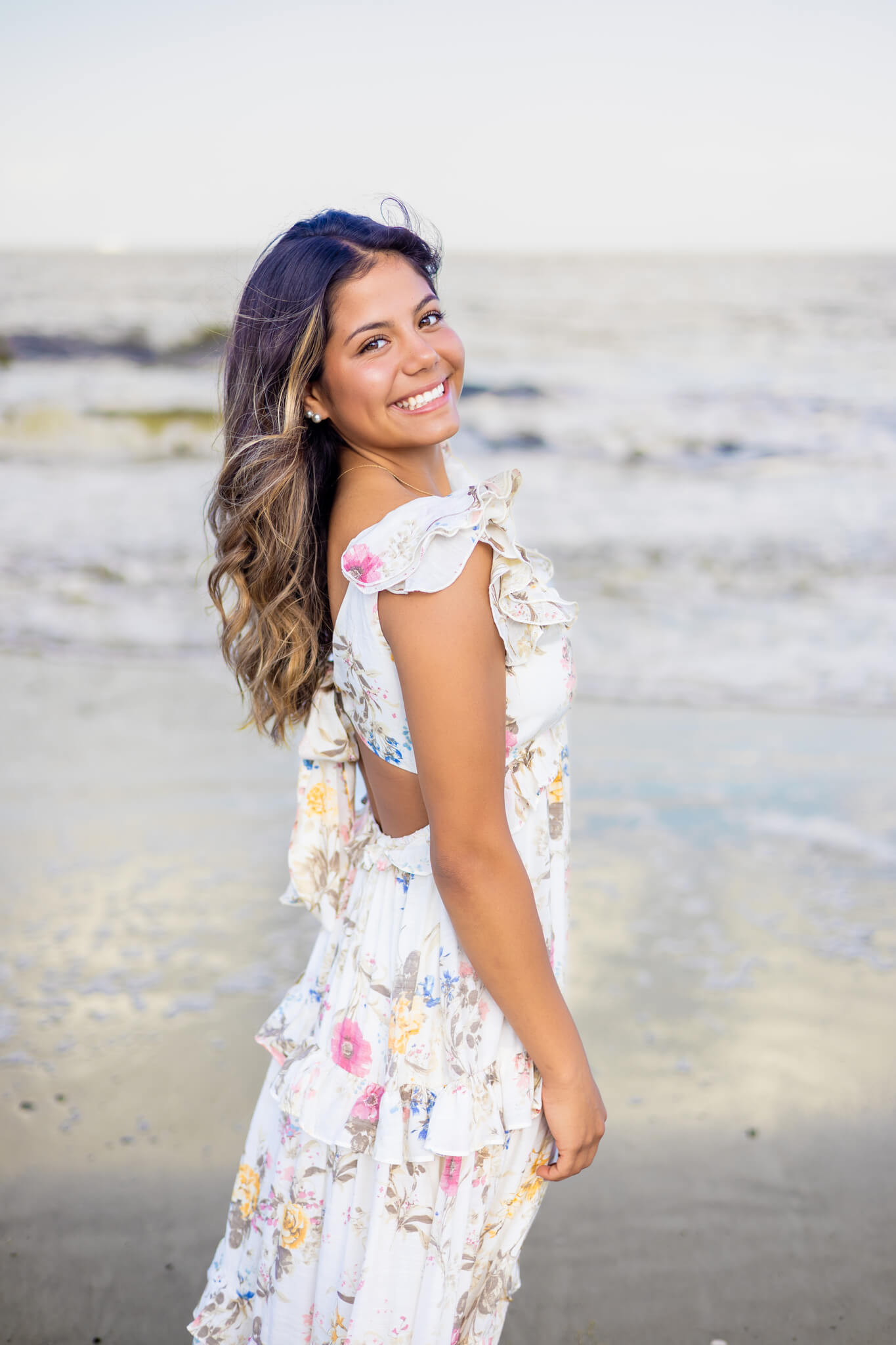 A happy high school senior with flowing hair and long flower print dress smiles over her shoulder while walking on a beach at sunset