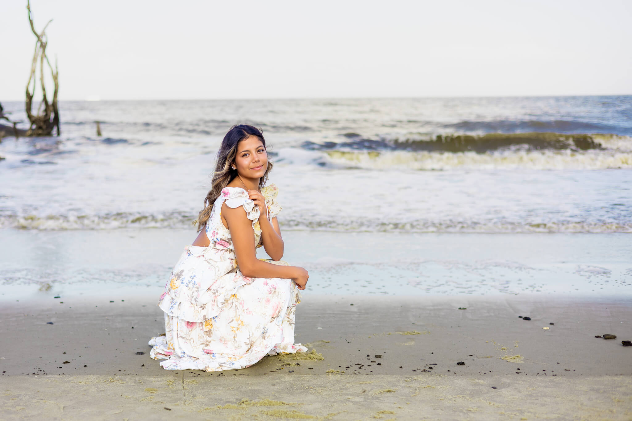 A high school senior in a white floral print dress kneels near the water on a beach at sunset smiling after exploring event spaces in richmond hill ga