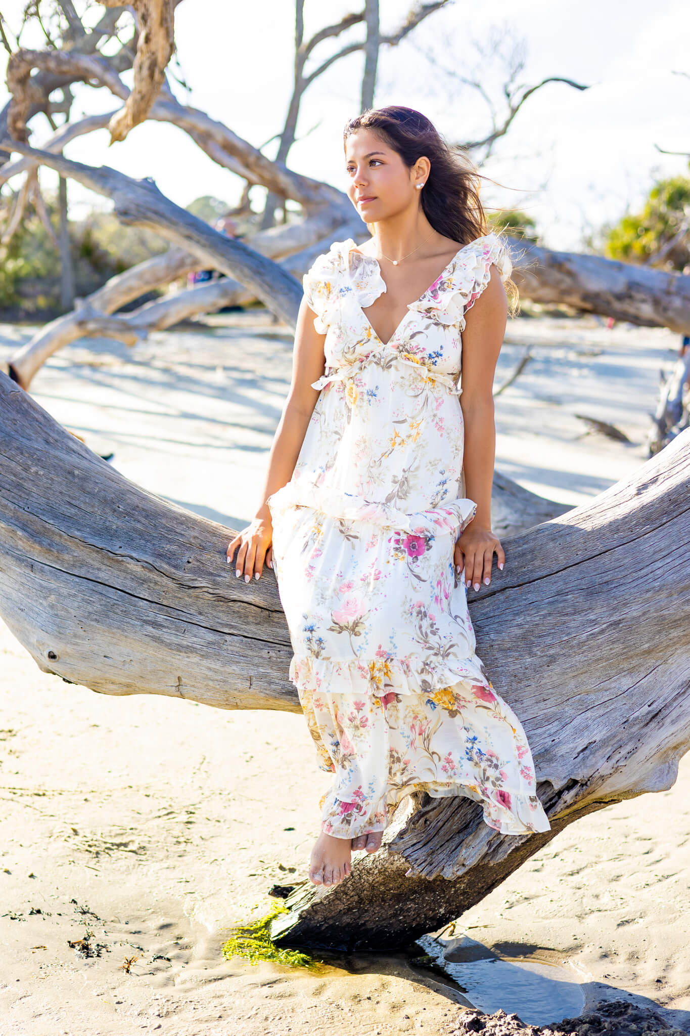A high school senior in a white floral print dress sits on driftwood on the beach after exploring event spaces in richmond hill ga