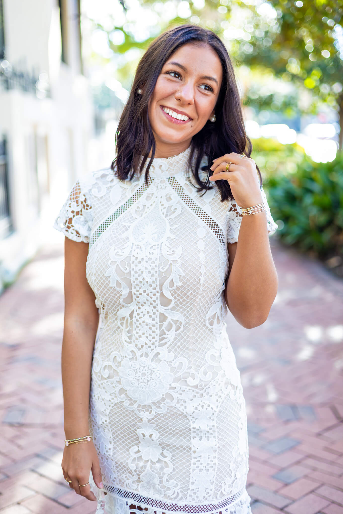 A happy woman in a white lace gown twirls her hair while walking a brick sidewalk