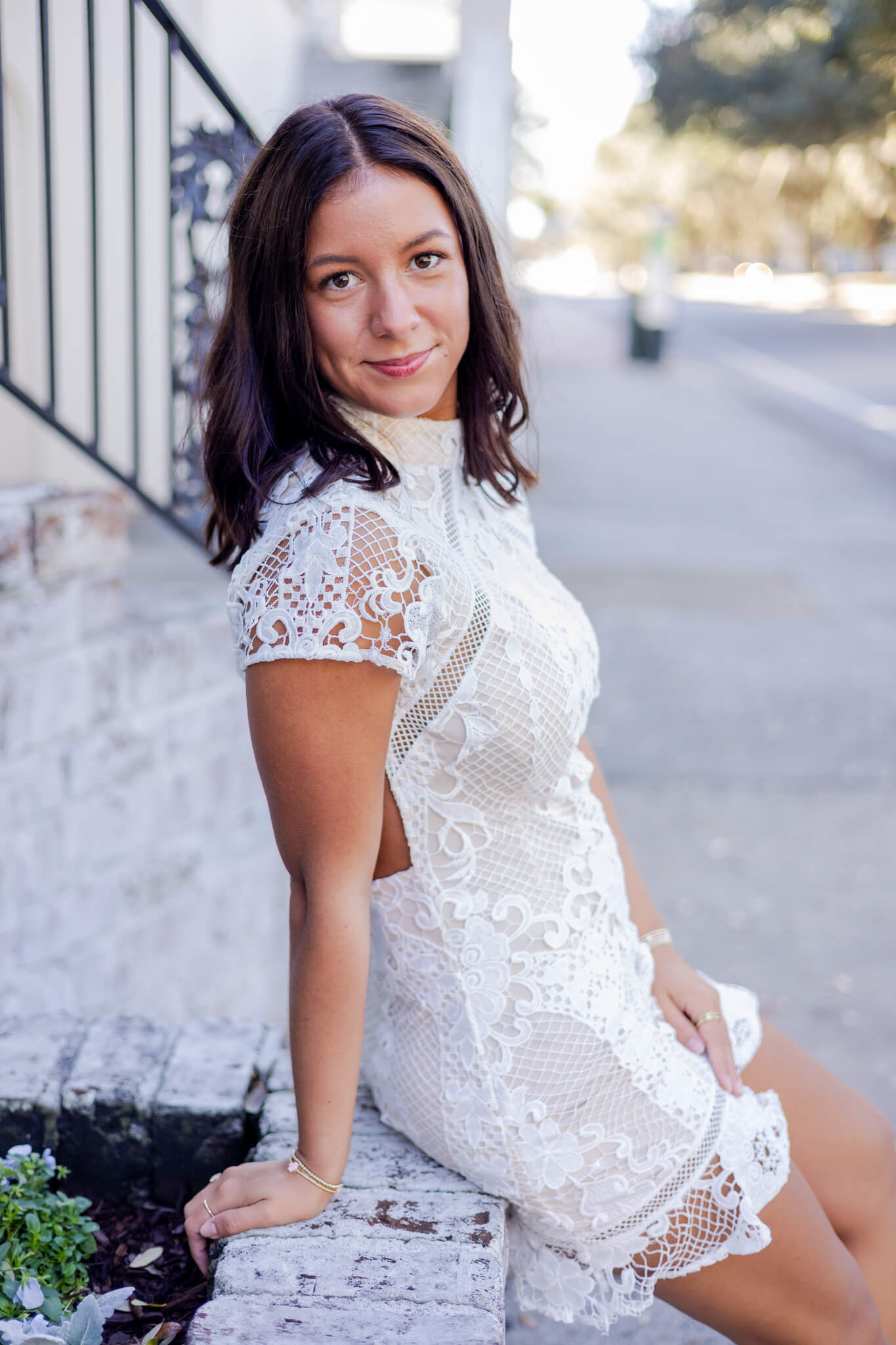 A smiling high school senior sits on a garden wall in a white lace dress after finding prom dresses in brunswick ga