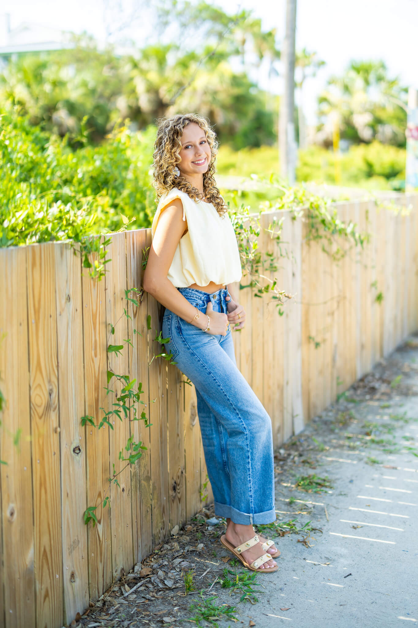 A smiling high school senior in a yellow blouse and jeans leans against a fence