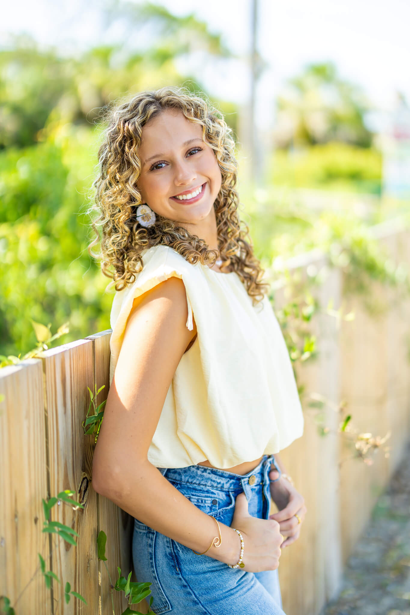 A smiling high school senior in a yellow blouse and jeans leans on a fence smiling with thumbs in belt loops after finding tanning in richmond hill ga