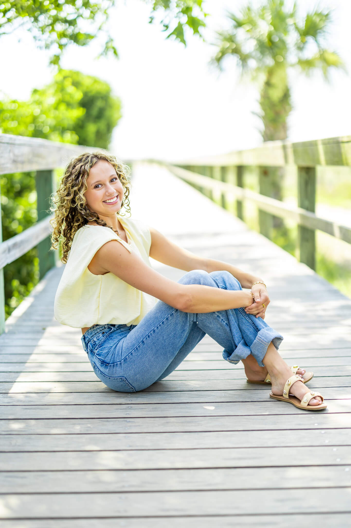 A happy high school senior sits in the middle of a boardwalk in jeans and yellow blouse after finding tanning in richmond hill ga