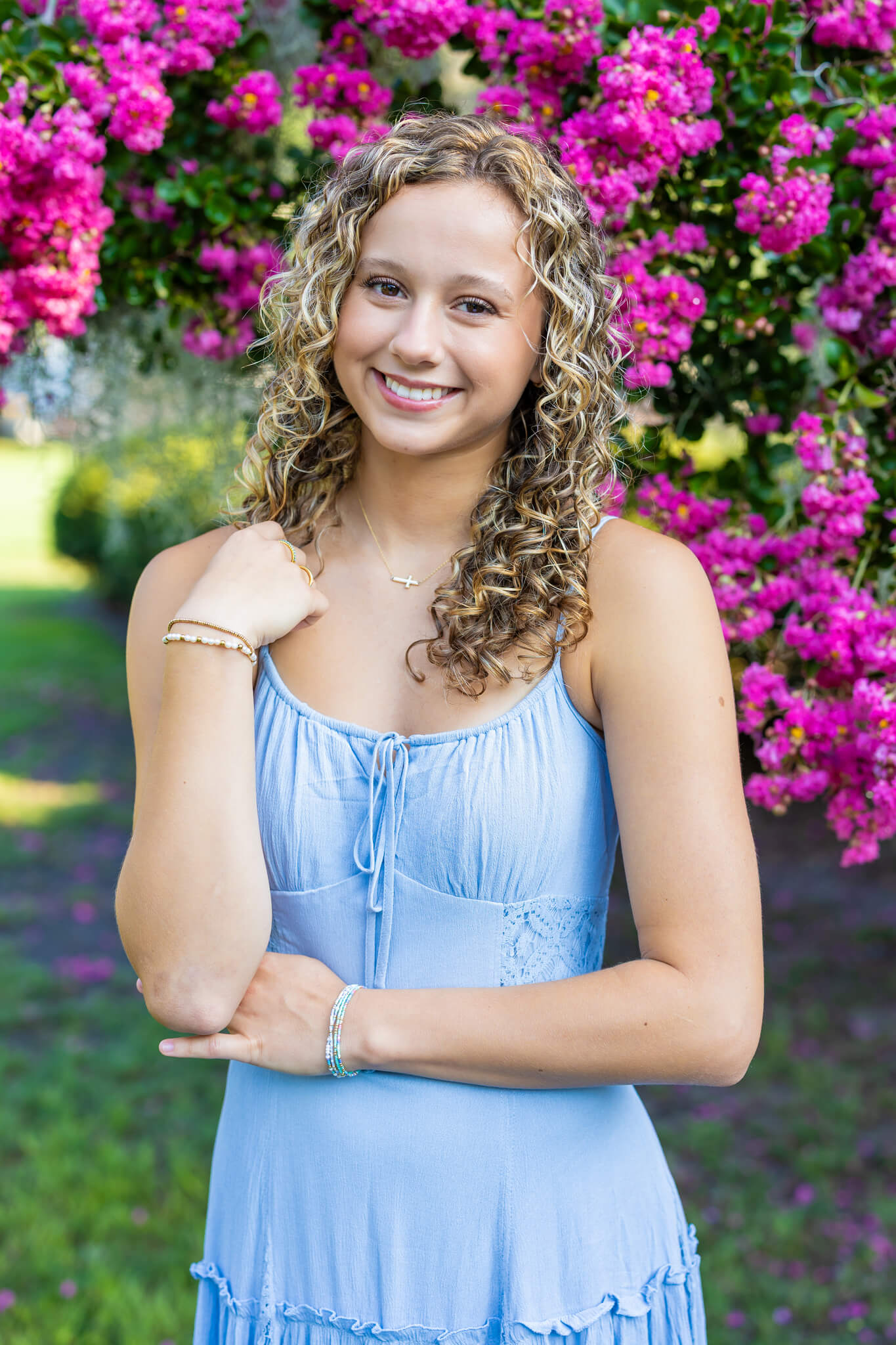 A smiling high school senior in a blue dress stands under a pink tree in a park after visiting that dress