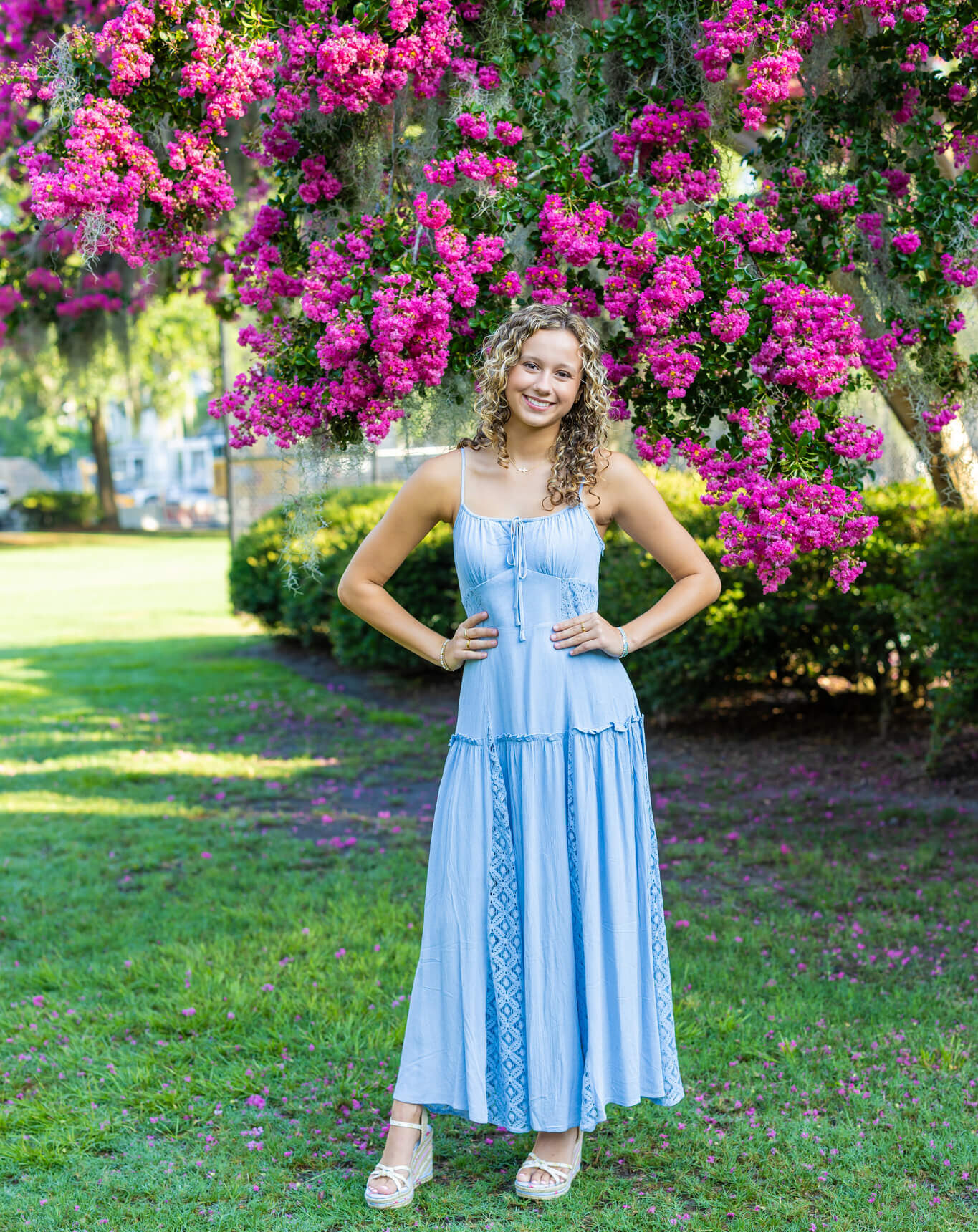 A smiling woman in a blue dress stands with hands on her hips under a flowering tree in a park after visiting that dress